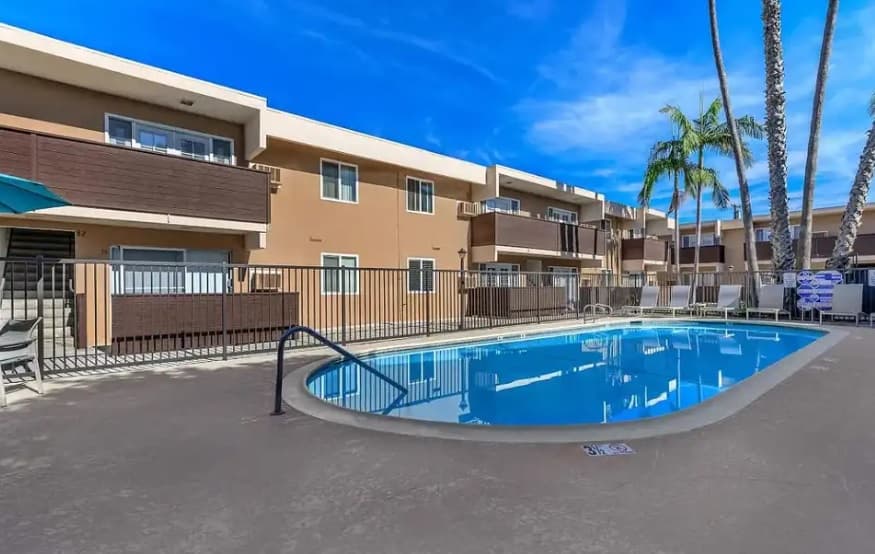 A clear blue swimming pool at Dorado Plaza is surrounded by a concrete deck and lounge chairs, with a black metal fence and two-story tan apartment buildings in the background under a bright blue sky dotted with palm trees.