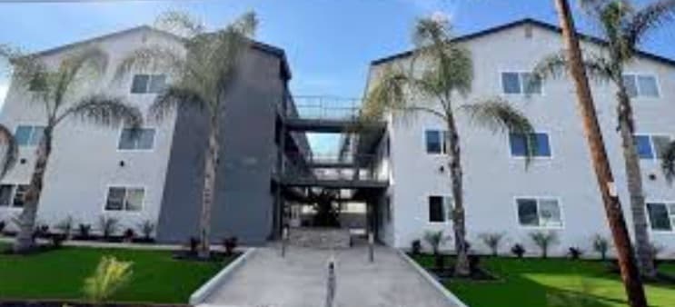 Two modern, white apartment buildings with palm trees in front, separated by a walkway and connected by open-air corridors, under a blue sky.