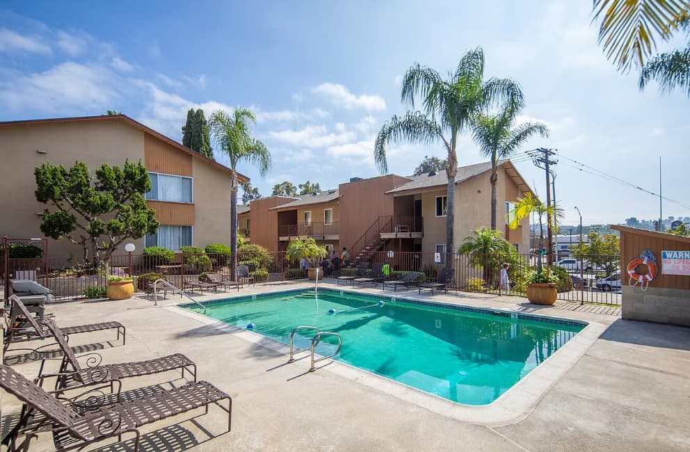 Sunny outdoor pool area at Fletcher apartments, surrounded by lounge chairs, potted plants, and palm trees, with two-story tan buildings in the background and a partly cloudy sky above.