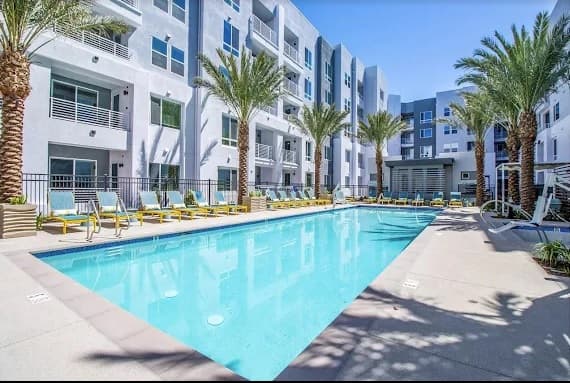 Modern apartment complex with several stories surrounding a rectangular outdoor swimming pool, lined with lounge chairs and palm trees under a bright blue sky.