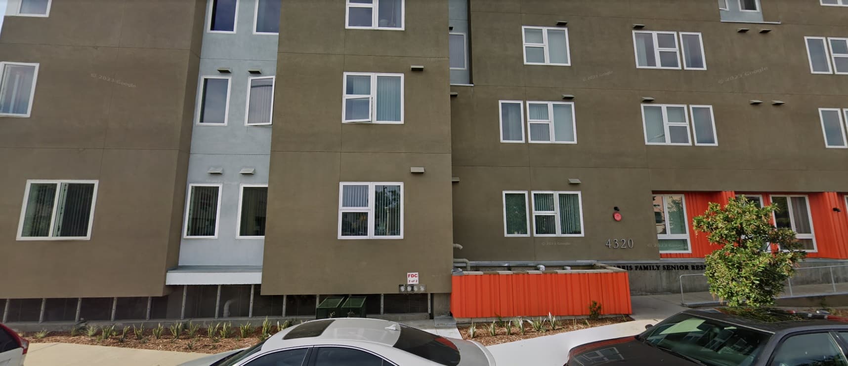 A modern senior residence with green and gray walls, white-framed windows, an orange accent panel, and parked cars along the street in front—the Harris Family Senior Residence offers stylish senior living.