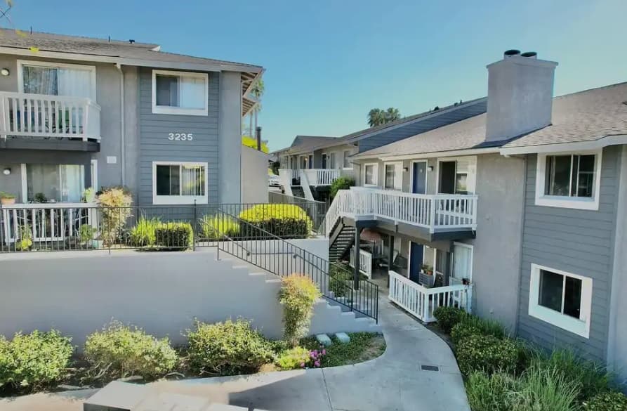 Hillside Terrace Apartments features two-story gray buildings with white trim and balconies, nestled among greenery and landscaping, all connected by a curved sidewalk and outdoor stairways on a sunny day.