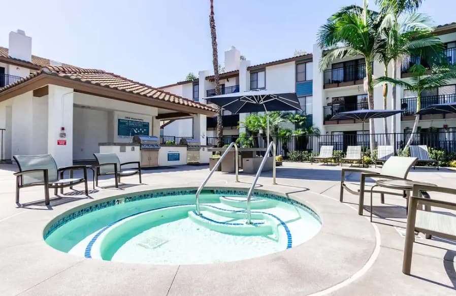 A round outdoor hot tub with steps is surrounded by lounge chairs and umbrellas in a sunny courtyard at IMT Mission Valley, with palm trees and modern luxury apartments in the background.