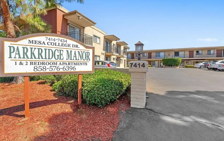 Exterior view of Parkridge Manor apartment complex with a sign displaying the address, phone number, and available 1 & 2 bedroom apartments. The building, parking area, and landscaping are visible.