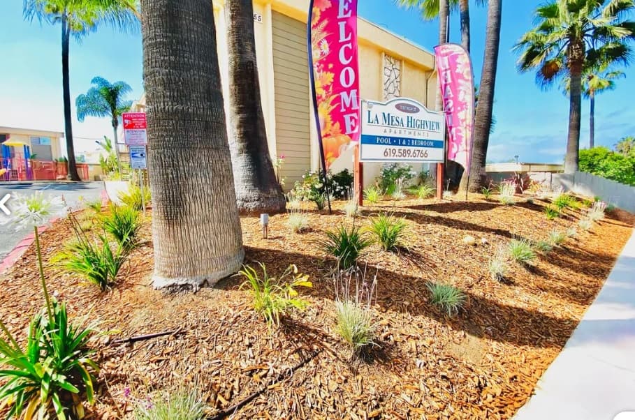 A landscaped area with palm trees and mulch surrounds a sign for Highview Apartments. Colorful flags and a “Welcome” banner are displayed near the building, inviting you to discover your new home in La Mesa under a bright blue sky.