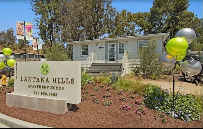 Single-story building with stairs, surrounded by trees and landscaping. A sign in front reads Lantana Hills Apartment Homes with a phone number while colorful balloons decorate the entrance to these inviting apartments.