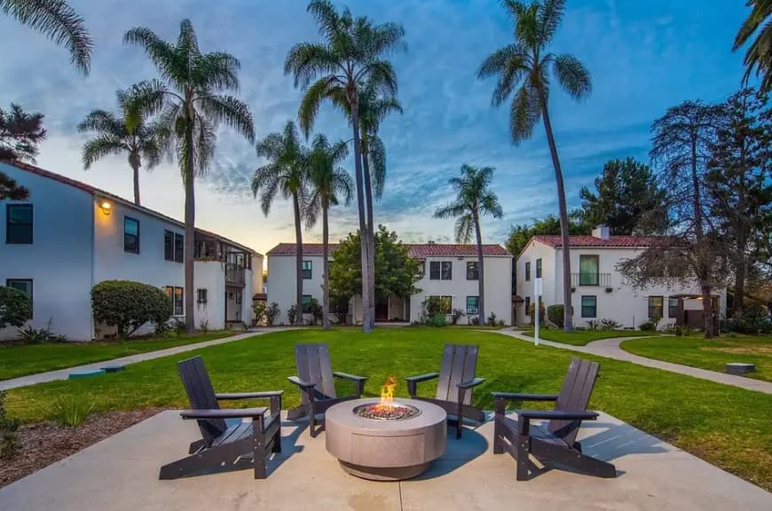 A circular fire pit surrounded by five dark chairs sits on a patio at Las Palmas Apartments, with palm trees and white two-story buildings in the background under a blue evening sky.