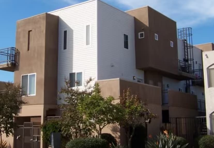 Modern luxury apartment building on Lauretta Street with a boxy design, white and brown exterior walls, multiple balconies, and lush trees and plants beneath a blue sky.