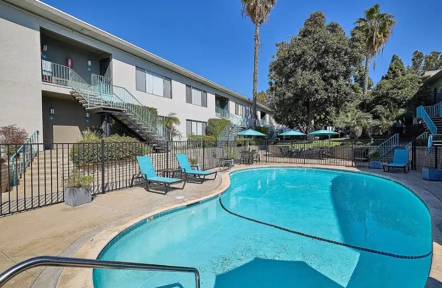A kidney-shaped outdoor swimming pool surrounded by lounge chairs, umbrellas, and two-story Mission Arbor Apartments with exterior staircases under a clear blue sky.