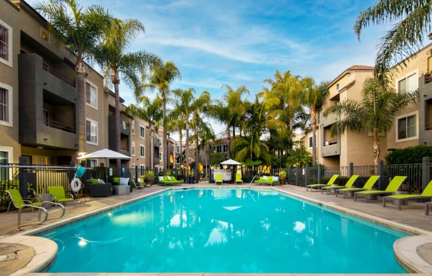 Outdoor swimming pool at Mission Pacific Apartments surrounded by palm trees, lounge chairs, and apartment buildings under a blue sky with scattered clouds. Poolside umbrellas and lush greenery create a relaxing atmosphere.
