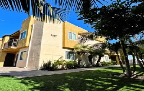 Morley-18 Apartments is a two-story yellow and beige building with several windows and small balconies, surrounded by palm trees and green lawn, under a clear blue sky.