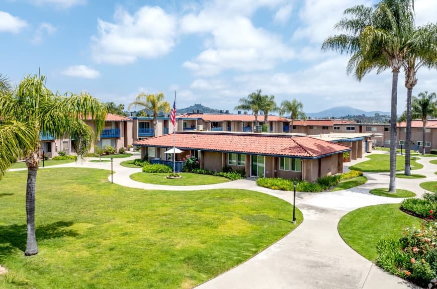 A landscaped courtyard at Lake Murray Villa features curved walkways, green lawns, palm trees, and single-story buildings with red-tiled roofs under a partly cloudy sky. An American flag stands near the center of these inviting villa rentals by Lake Murray.