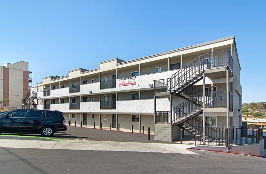 A three-story Pacific Cove apartment building with exterior walkways, a metal spiral staircase, and a large black SUV parked in front under a clear blue sky. A “Now Leasing” sign is displayed on the second floor railing.