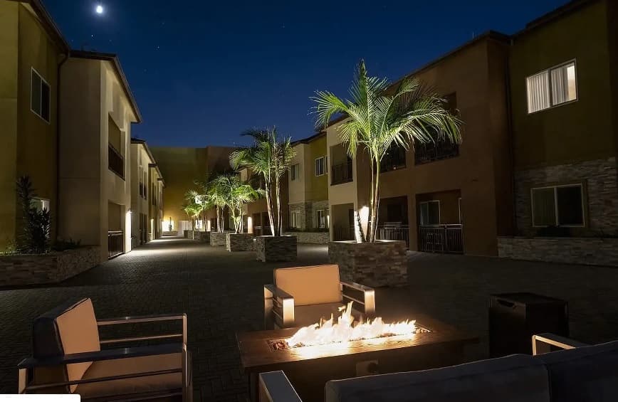 A cozy outdoor patio at night features a fire pit surrounded by four cushioned chairs. Palm trees and the softly lit Pacific Heights Apartments line the walkway in the background under a starry sky.