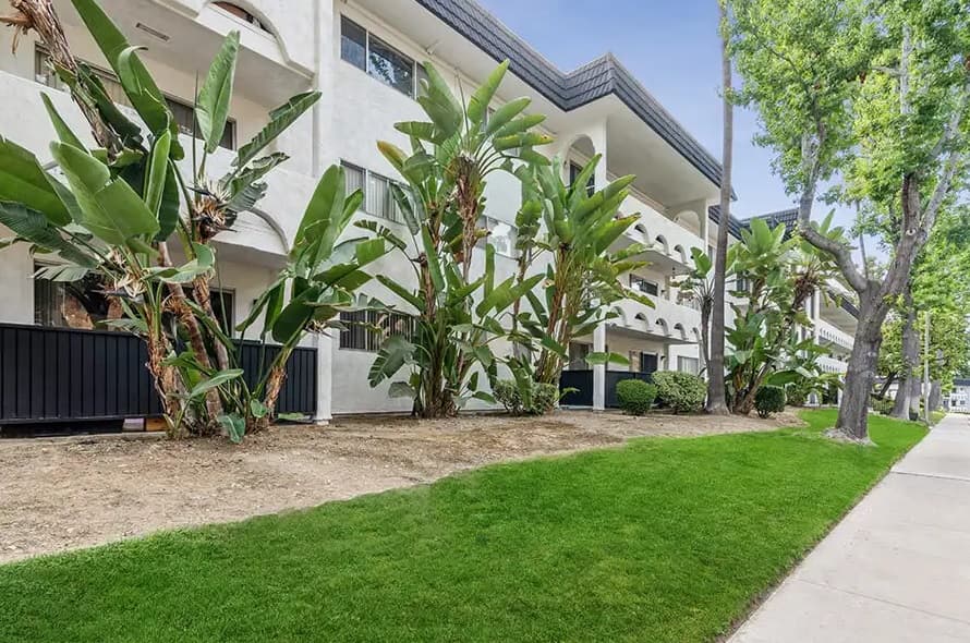 A modern white apartment building with balconies is bordered by lush green grass, tropical plants, and tall palm trees along a clean sidewalk under a clear blue sky.