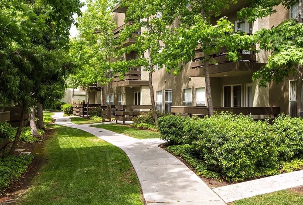 A curved sidewalk winds through Parkview Terrace, a landscaped apartment complex with green grass, bushes, leafy trees, and brown two-story buildings with balconies.
