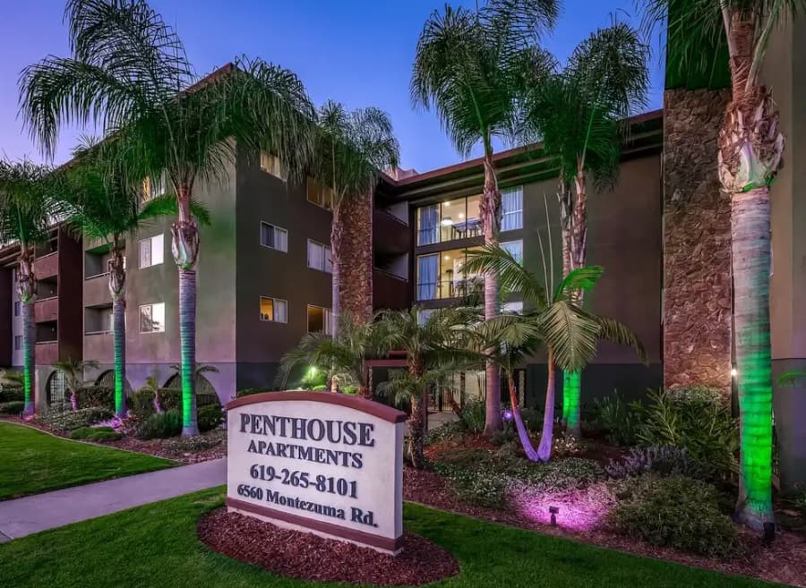 Exterior view of luxury penthouse apartments at dusk, featuring palm trees, green lawn, and a lit-up sign with the 6560 Montezuma Rd address and phone number, set before a multi-story building for truly upscale living.
