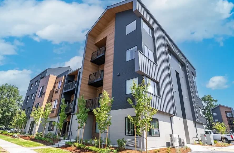 Modern three-story apartments in Grand Forks, The Ponds on Alexander feature wood and dark gray siding, balconies, large windows, and young trees lining the sidewalk under a bright blue sky with scattered clouds.