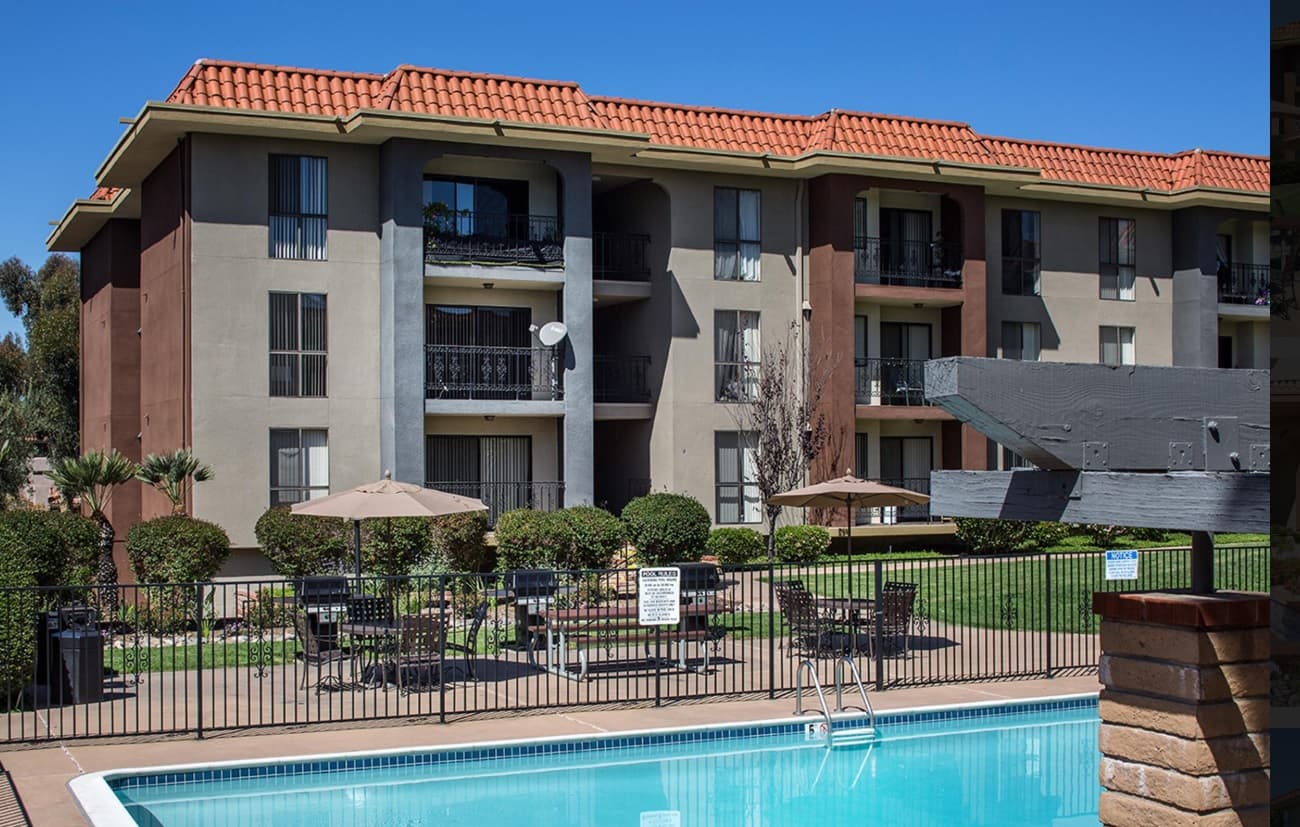 Prado Apartments is a three-story rental property with a red tiled roof, balconies, and a fenced-in outdoor pool with lounge chairs, tables with umbrellas, and lush landscaping under a clear blue sky.