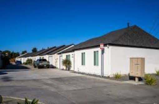 A row of renovated villas with dark roofs lines a clean driveway on a sunny day. A truck is parked outside, and a beige mailbox stands near the curb. Each villa features a private EV hookup under the clear blue sky.