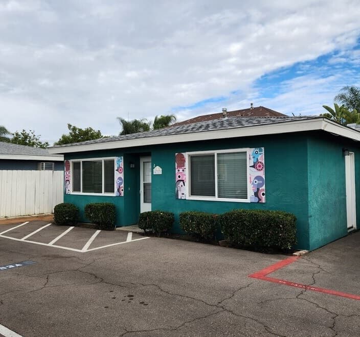 A small, teal one-story building at Rolando Place Apartments with two large windows, colorful banners by the entrance, trimmed bushes out front, and a parking area with handicap spaces. The sky is mostly cloudy.