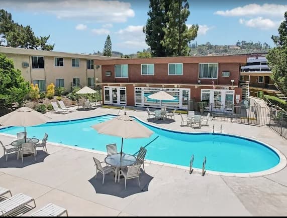 A fenced outdoor swimming pool at Stadium Arms Apartments is surrounded by lounge chairs, tables with umbrellas, and apartment buildings in the background, all basking in the sun.