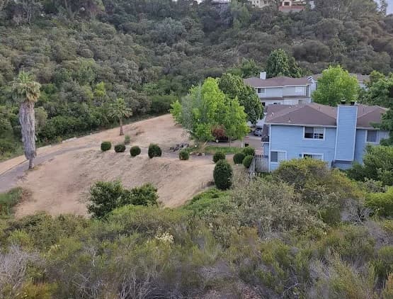 Row of light blue houses bordered by trees and shrubs, with a dirt path and dry grassy area in front, nestled at the edge of a wooded hillside near Talmadge Canyon Park.