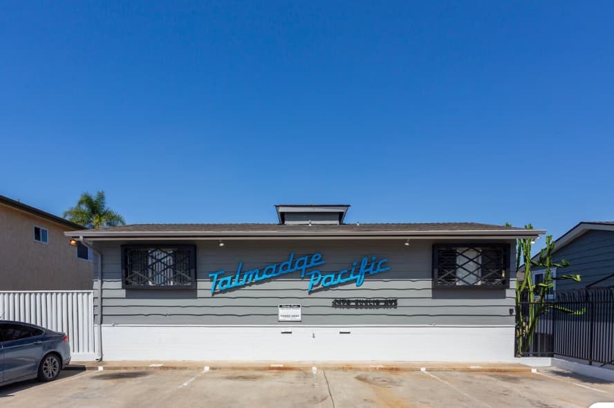 A gray, single-story building with barred windows features a blue neon sign reading Talmadge Pacific Apartments against a clear blue sky. A car is partially visible on the left, and a white fence and gate are on either side.