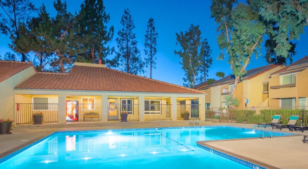 A brightly lit outdoor swimming pool is surrounded by lounge chairs and bordered by two beige buildings with red-tiled roofs and large windows at Terra at Mission Trails, set against tall pine trees at dusk.