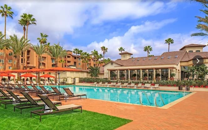 Relax at The Village Mission Valley’s outdoor swimming pool, featuring lounge chairs and orange umbrellas, surrounded by palm trees and a large building with a tiled roof under a partly cloudy sky.