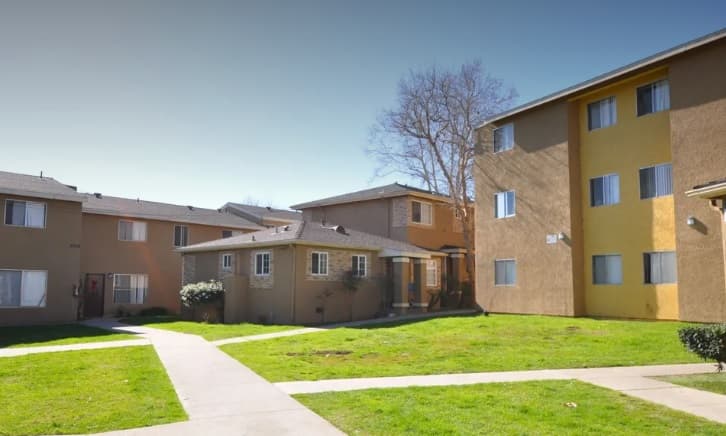 A courtyard view of a residential apartment complex with tan and yellow buildings, green grass, and paved walkways on a clear, sunny day.