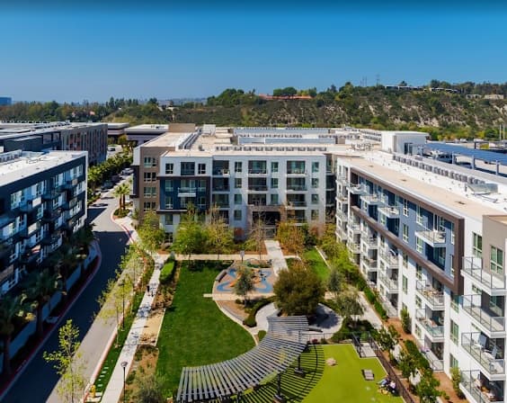 A modern apartment complex with several multi-story buildings surrounds a landscaped courtyard featuring green lawns, trees, benches, and a small playground, set against a backdrop of hills under a clear blue sky.