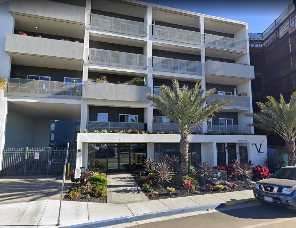 A modern four-story apartment building with balconies, palm trees, and landscaped plants in front. A black car is parked at the curb, and the building has a white facade with V. signage near the entrance.
