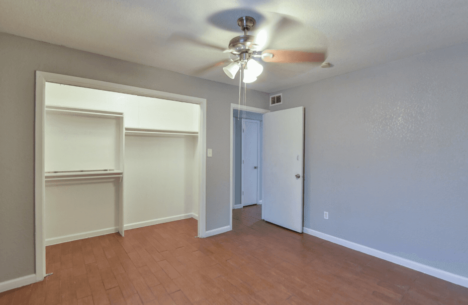 Empty bedroom with light gray walls, wood floor, ceiling fan with lights, open closet with shelves and rods, and a white door partially open, revealing a hallway.