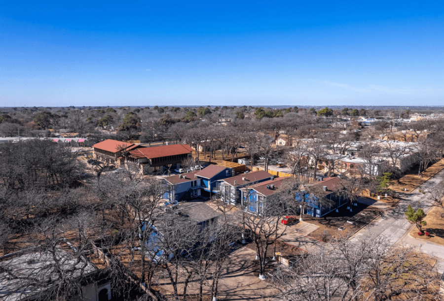 Aerial view of a suburban neighborhood with blue apartment buildings and leafless trees, under a clear blue sky; roads and other buildings are visible in the background.