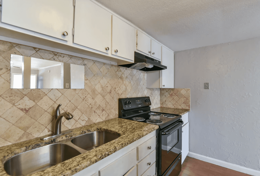 A small kitchen with a granite countertop, stainless steel double sink, electric stove, oven, white cabinets, and beige tiled backsplash. A slim mirror is mounted above the sink.