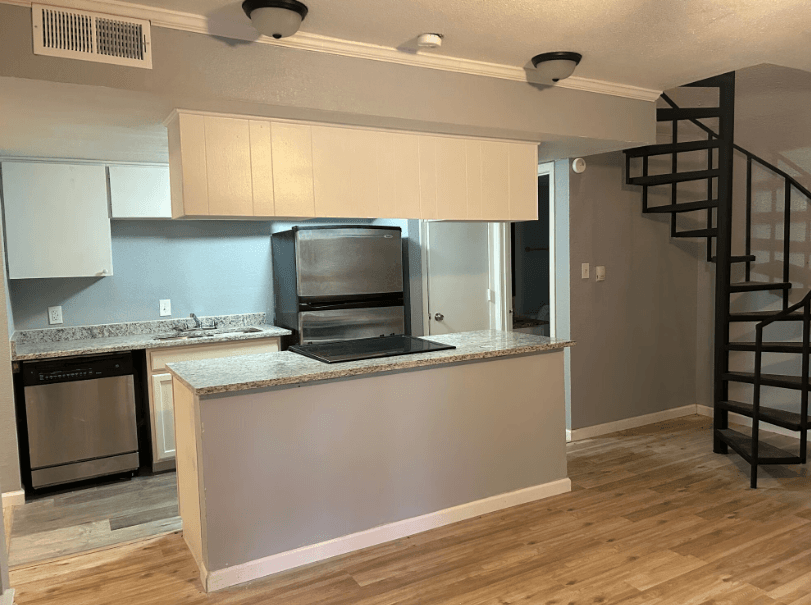 Modern kitchen with light wood floors, gray and white cabinets, granite countertops, stainless steel appliances, and a black spiral staircase leading upstairs. Walls are painted light gray with white trim.