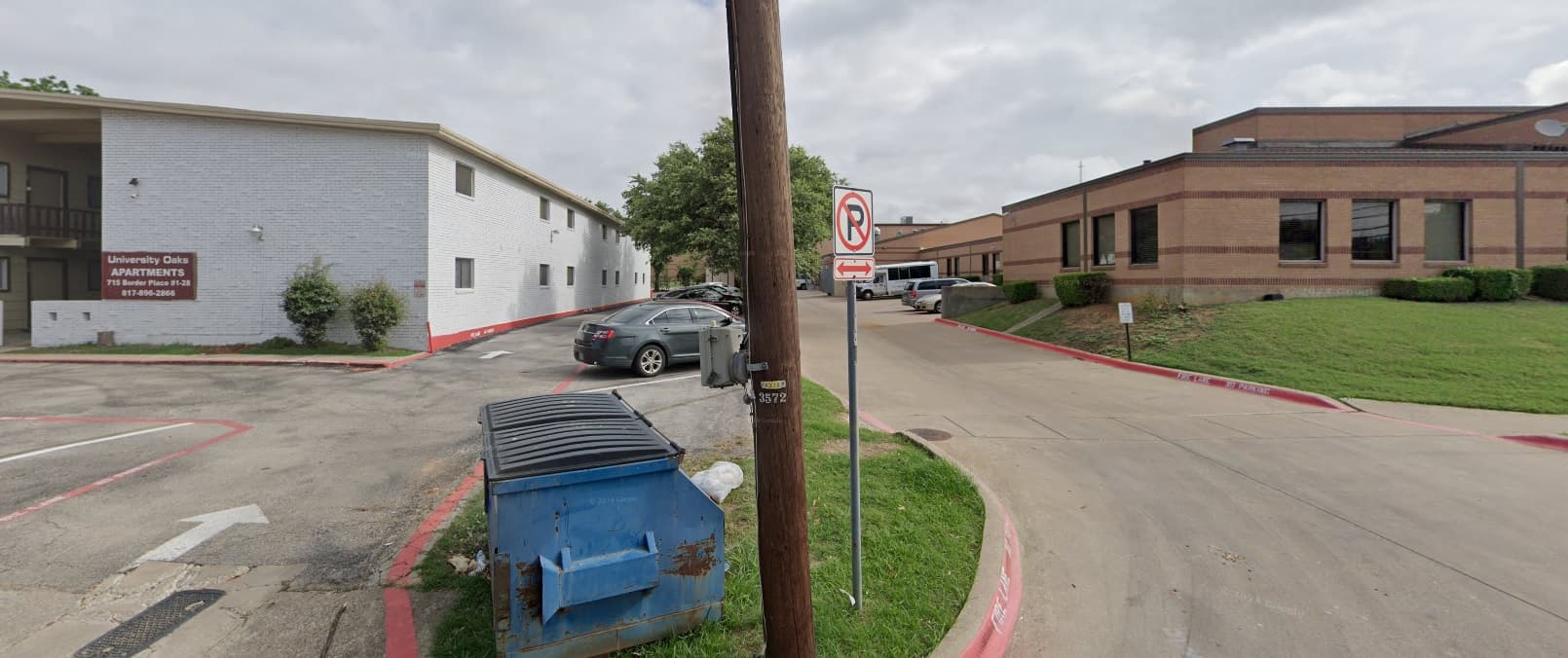 A no parking sign is posted on a wooden pole in front of a blue dumpster at University Oaks Apartments Arlington. To the left is a white brick apartment building, to the right a brown brick building, with cars parked along the driveway.