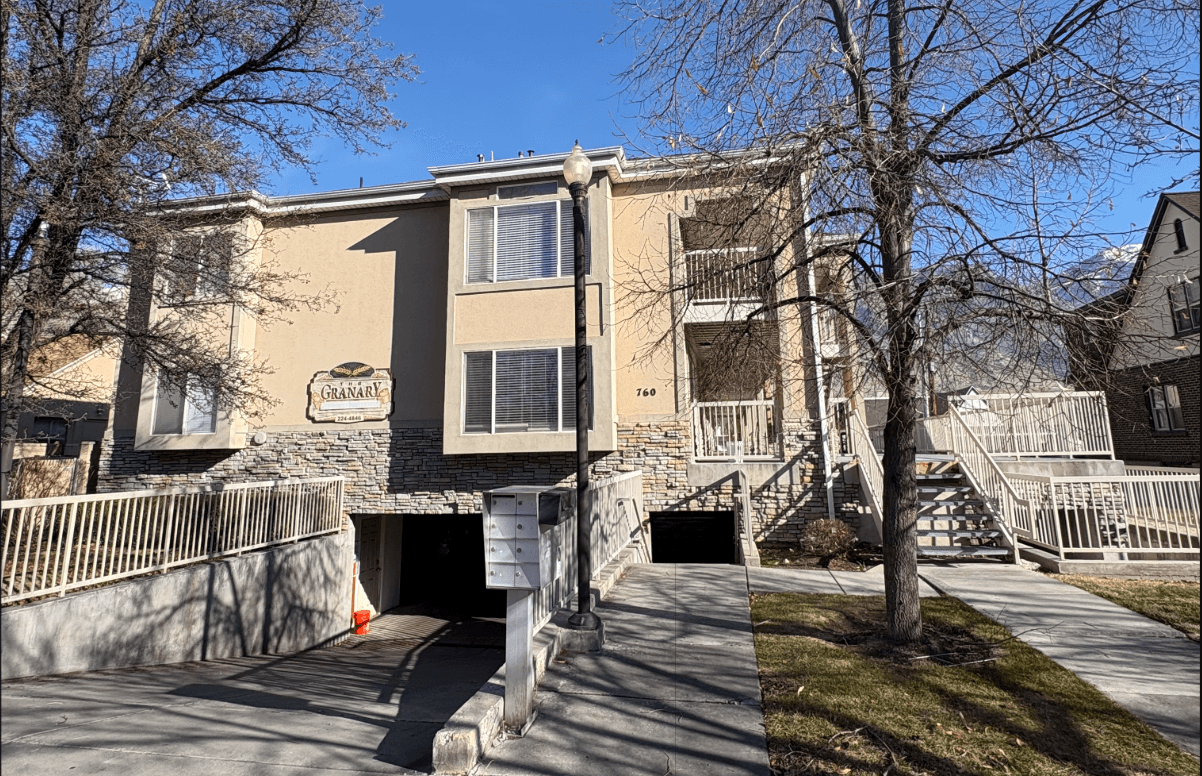 Granary Apartments is a two-story beige building with stone accents, two garage entrances, and white railings. Leafless trees and a row of mailboxes are visible out front beneath a clear blue sky.