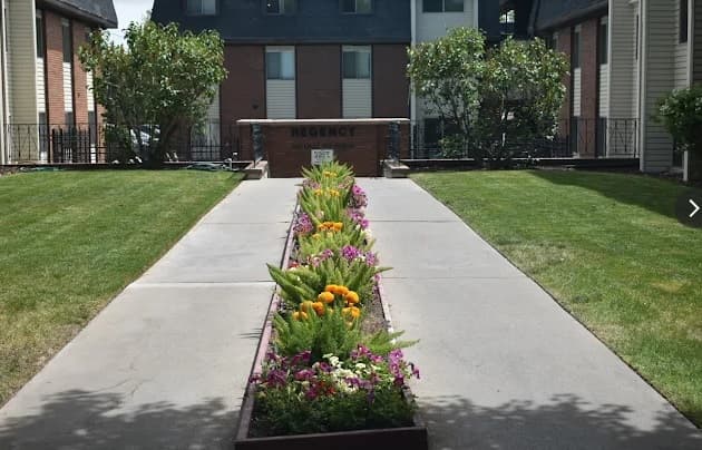 Concrete walkway flanked by green lawns and raised flower beds with colorful flowers, leading to a brick sign reading REGENCY in front of an apartment building.