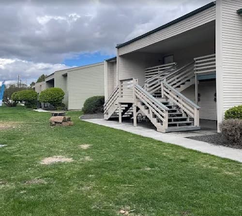 A beige apartment building on River Street features outdoor stairs, a small covered porch, and well-kept grass. Bushes line the sidewalk, and a picnic table rests on the lawn beneath a cloudy sky.