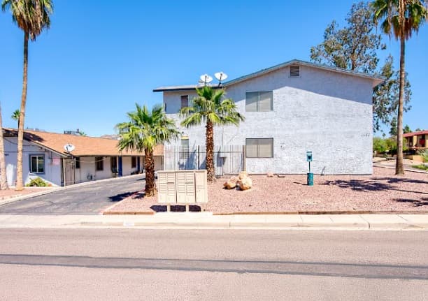 A two-story light gray building at 1452 E Cinnabar Ave with a sloped roof, palm trees, a mailbox cluster, and desert landscaping is showcased under the clear blue skies—an inviting example of Phoenix AZ real estate.