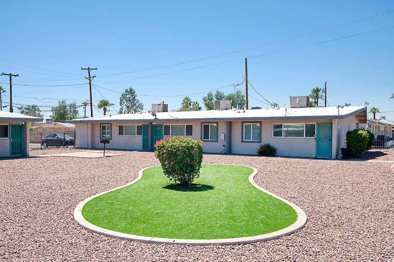 A single-story white brick house at 2201 W Heatherbrae Dr, Phoenix, AZ 85015, featuring barred windows, a metal security door, wooden gate, small garden area with sparse grass, and a clear blue sky in the background.