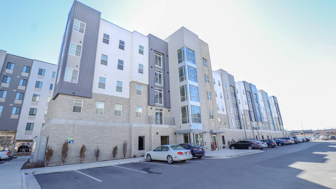A modern four-story apartment building, Promenade Place Orem features a gray and white exterior, large windows, and a blue balcony. Several cars are parked in front under a clear blue sky. The address 875 S is visible above the entrance.