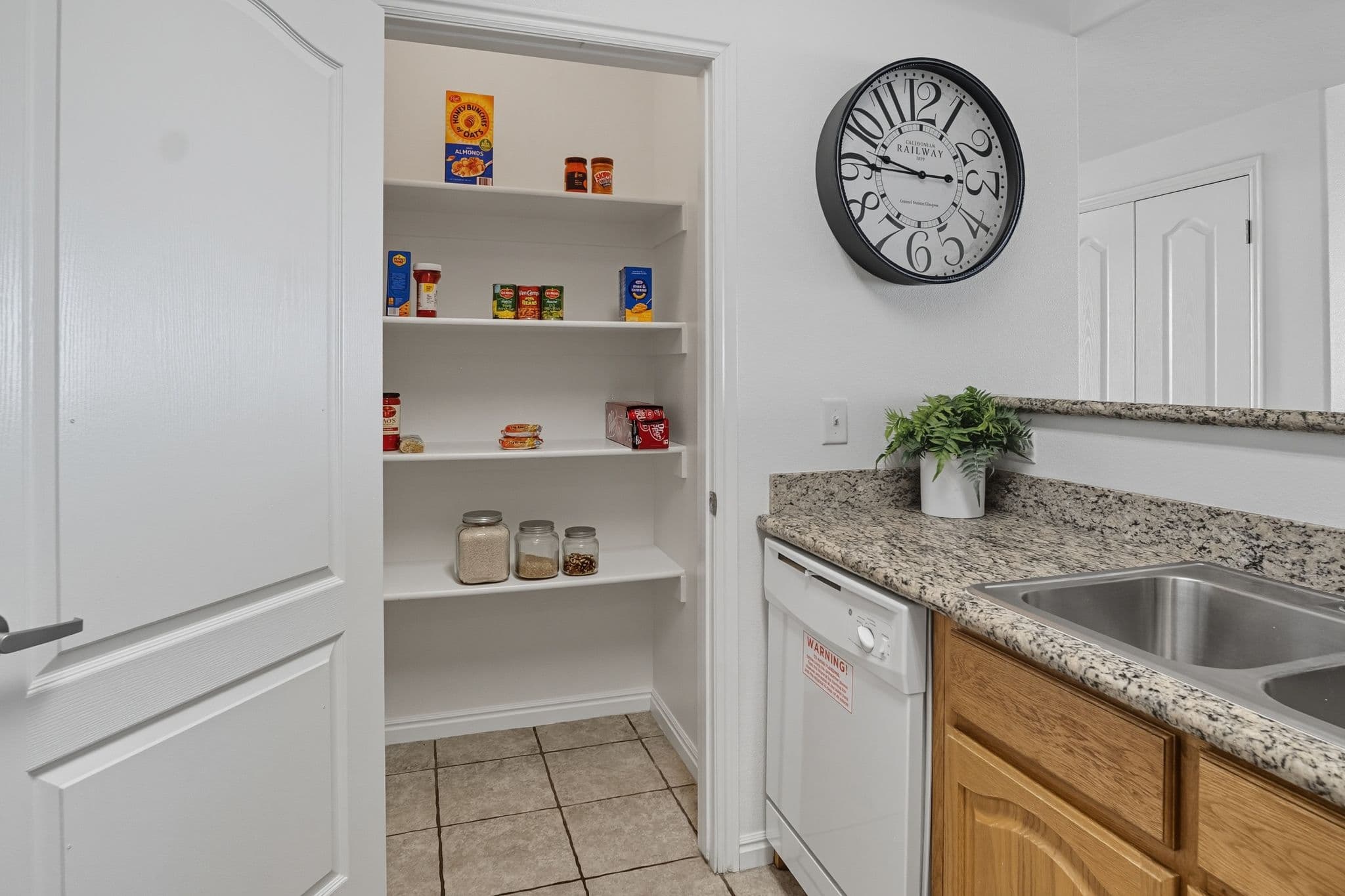 A kitchen at Ventana Student Housing Orem features granite countertops, a sink, and a dishwasher. Next to a large wall clock, an open pantry reveals shelves with jars and cans. A small plant decorates the counter.
