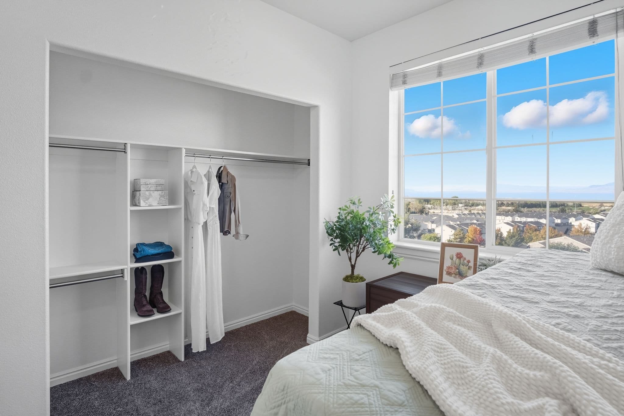 A bright, minimalist bedroom at Ventana Student Housing Orem features an open closet with dresses, shirts, boots, and shelves. A bed with a textured blanket sits beside a large window overlooking scenic views, with a plant and picture decorating the room.