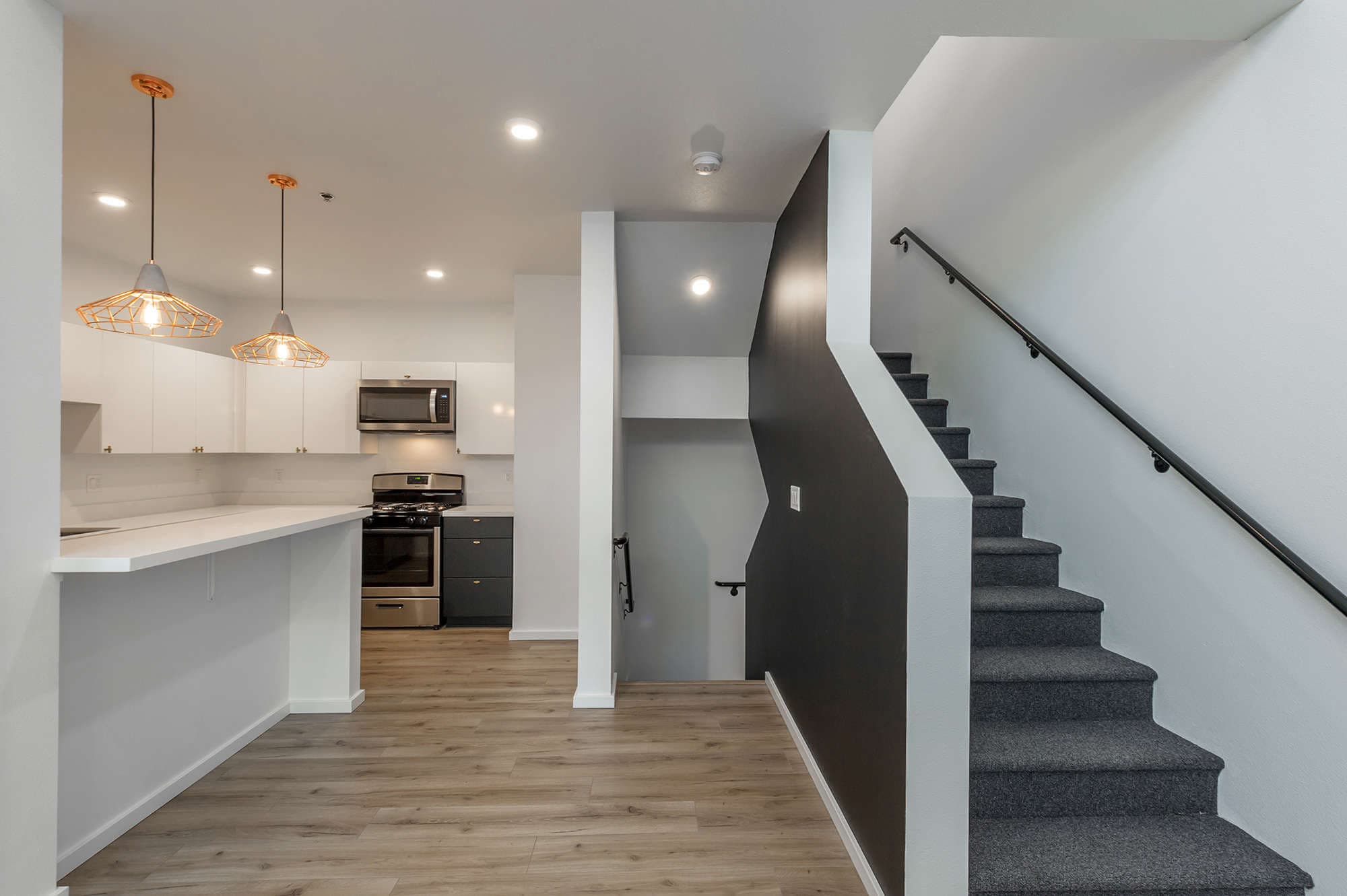Modern kitchen at The Urban Chico features white cabinets and stainless steel appliances beside a staircase with gray carpet and a black accent wall. Pendant lights illuminate the breakfast bar, and wood-look flooring completes the main area.