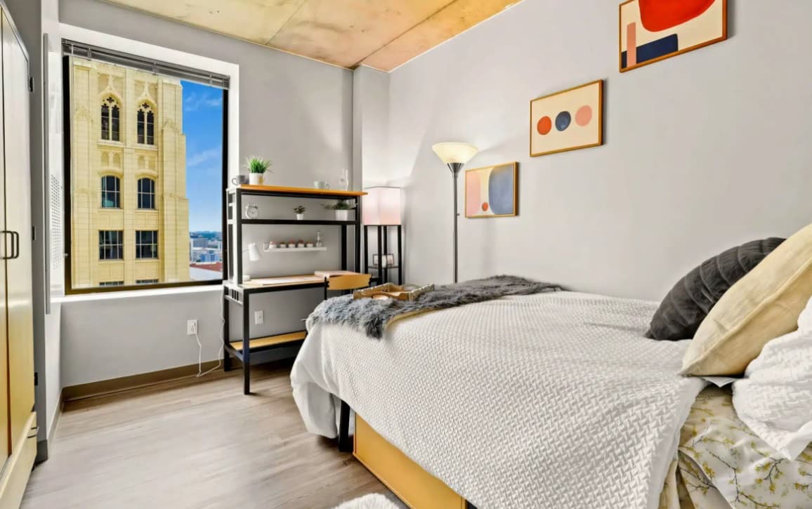 A modern bedroom at Auraria Lofts in Denver features a large window with a city view, a neatly made bed with gray and white bedding, a desk with shelves, a floor lamp, and abstract art on the light-colored walls—perfect for student housing.