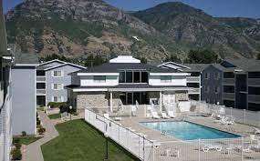 Carriage Cove Provo features multiple gray apartment buildings, a central clubhouse, and a fenced outdoor pool. Green lawns and walkways surround the complex, with mountains rising in the background under a clear blue sky.