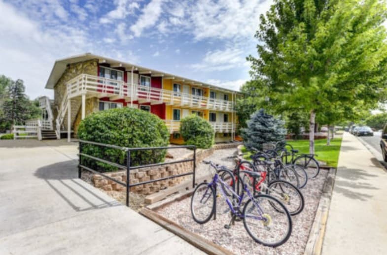 A row of colorful bicycles is parked in a bike rack outside Buffalo Canyon Apartments Boulder, a two-story building with white railings, surrounded by green bushes and trees under a partly cloudy sky.
